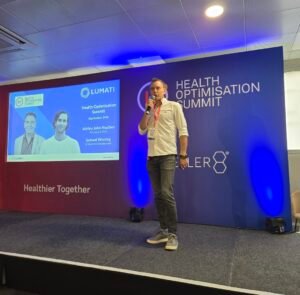 Ashley Heather speaking into a microphone on stage during a presentation at the Health Optimisation Summit.