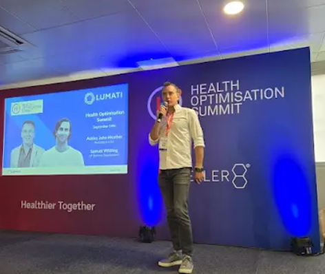 Ashley Heather holding a microphone and speaking on stage at the Health Optimisation Summit in front of a presentation screen.